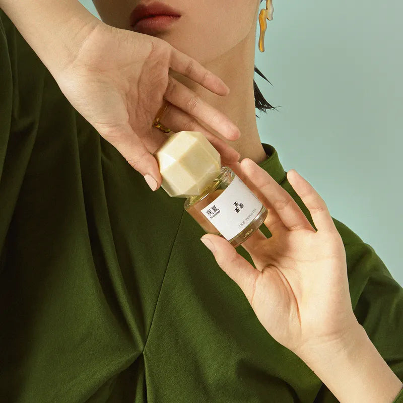 Model holding To Summer Triple Tea perfume with a beige cap, wearing green clothing against a soft teal background.